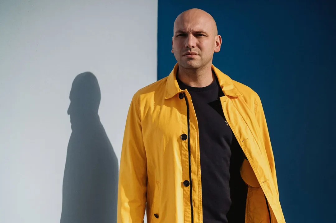 Esteban Fernandez en veste jaune devant un mur blanc et bleu, son ombre portée