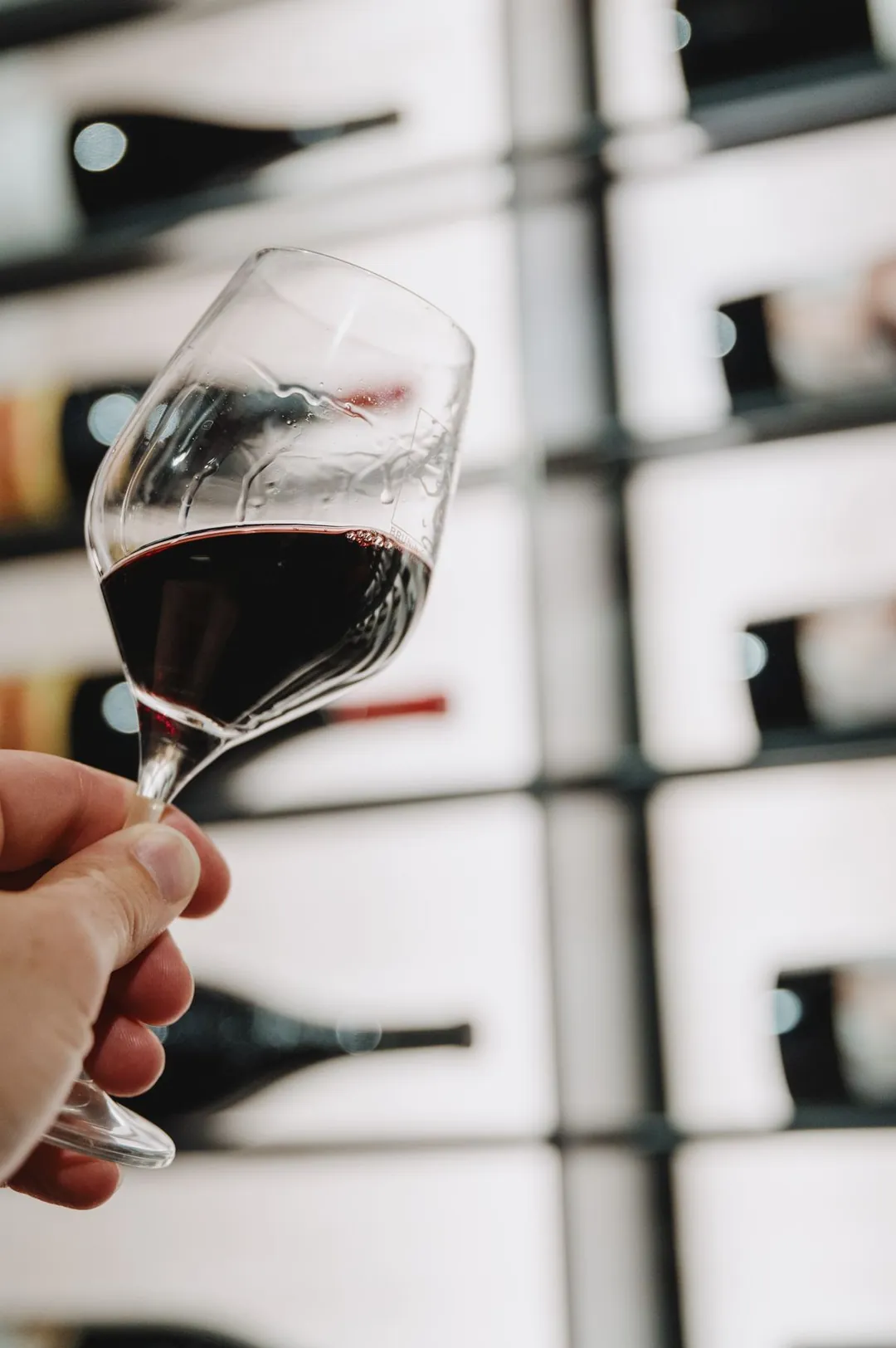 Main levant un verre de vin rouge devant des bouteilles
